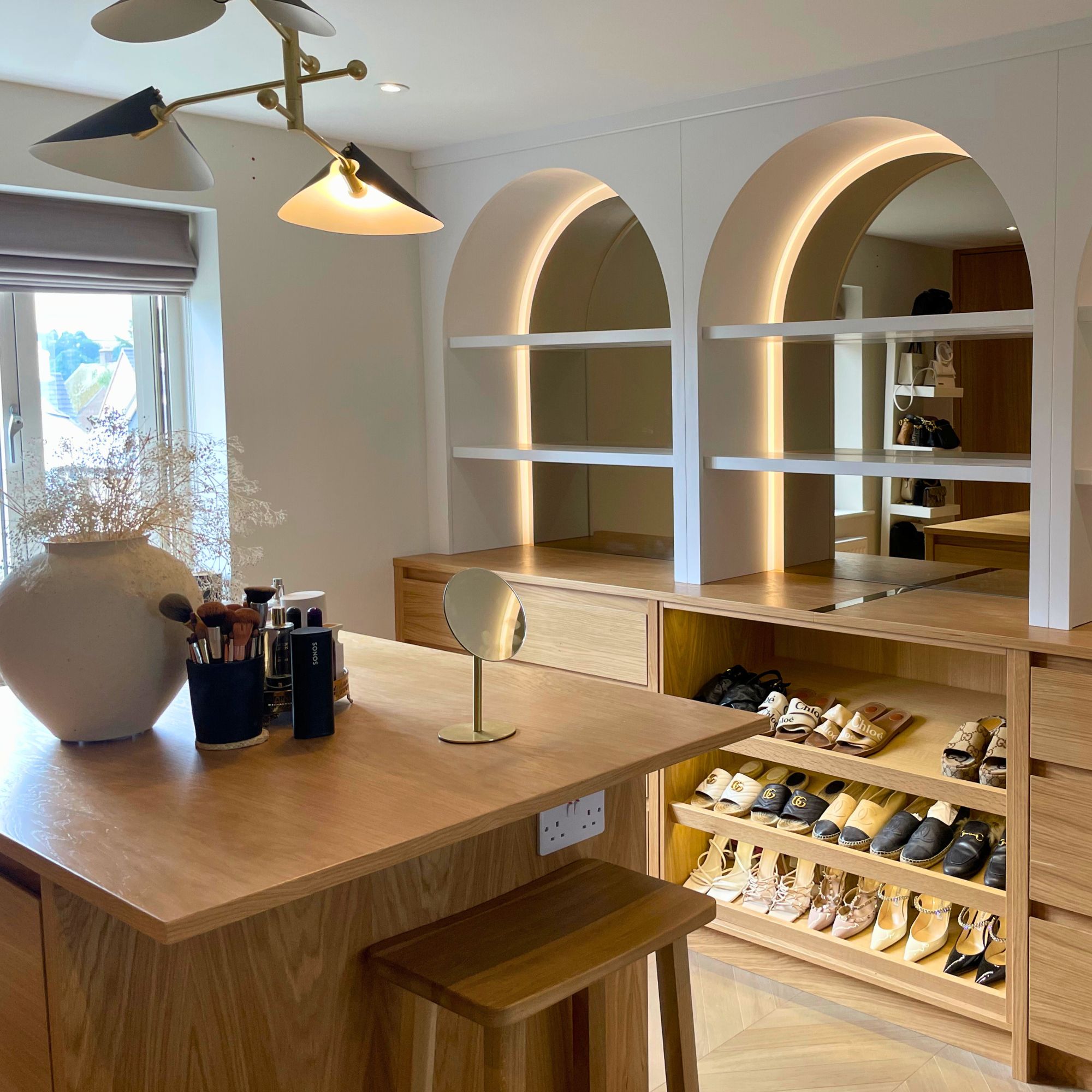 The image shows a custom domestic dressing room, featuring an island style table. In the background is a custom storage area featuring 6 drawers with a shoe rack in the middle, with a series of mirrored arches above