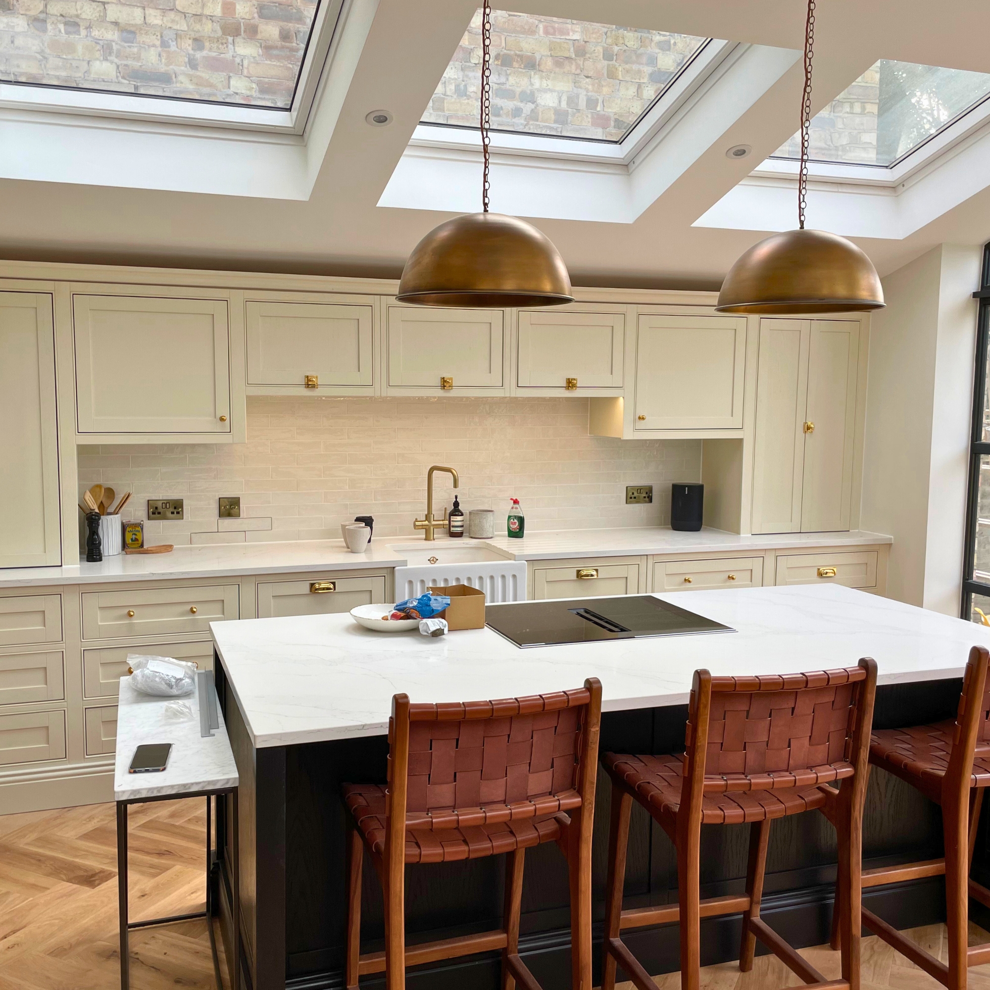 The image shows a bespoke kitchen. The kitchen is predominantly cream, with a black fronted island featuring 3 red stools. The kitchen has hanging brass lights next to 3 sky lights